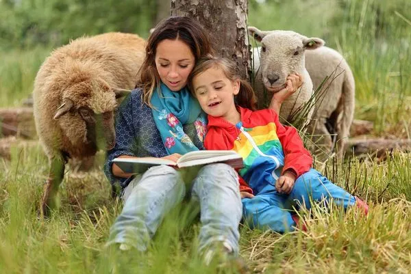 A young woman and a girl sit in long grass against a tree reading a book as two sheep look on around the tree.